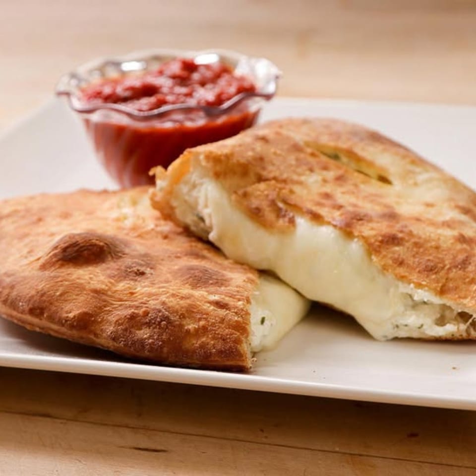 Two golden-brown calzones, one cut open to reveal melted cheese, with a side of red marinara sauce.