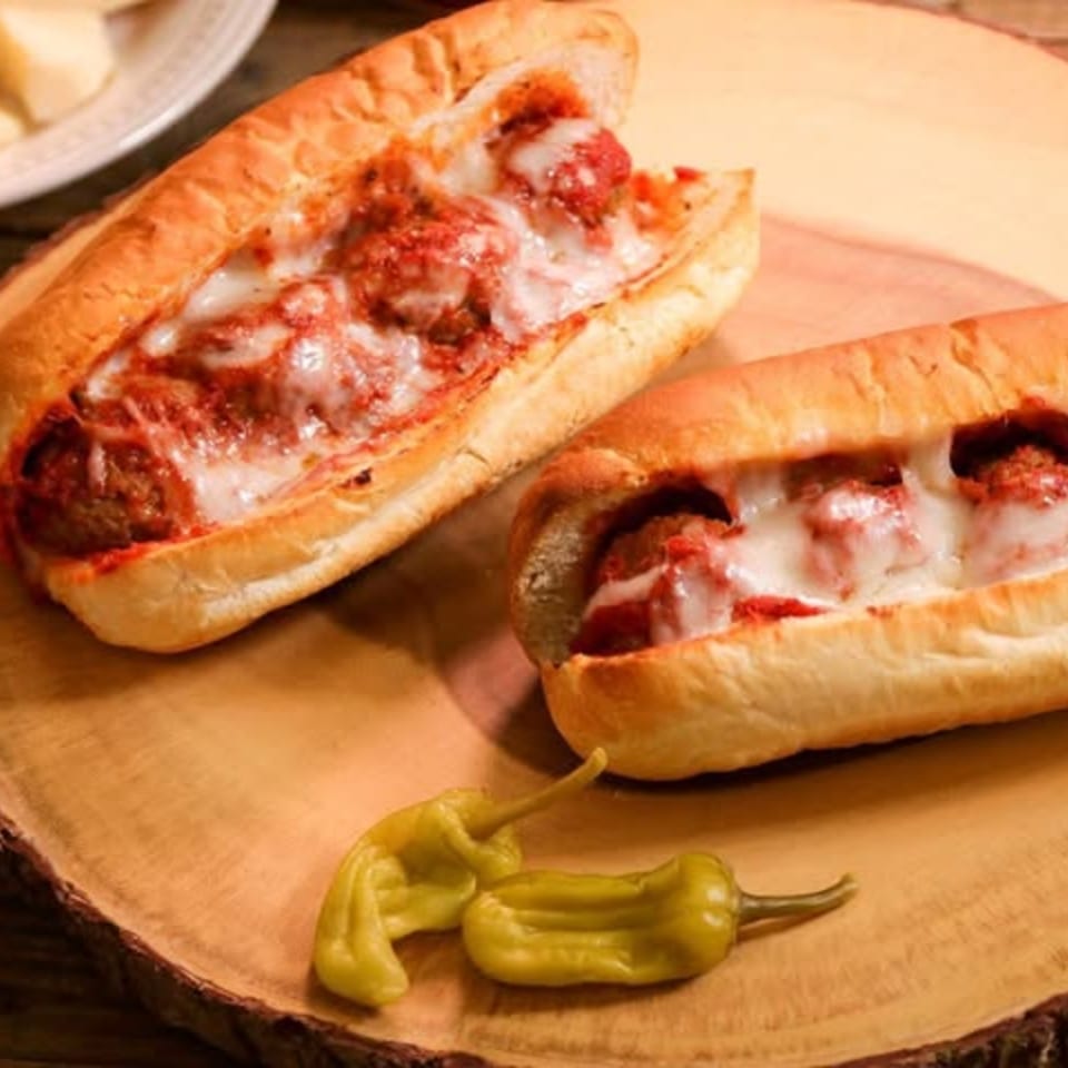 Two Ameci Pizza & Pasta meatball subs with melted cheese and sauce, garnished with two green peppers.